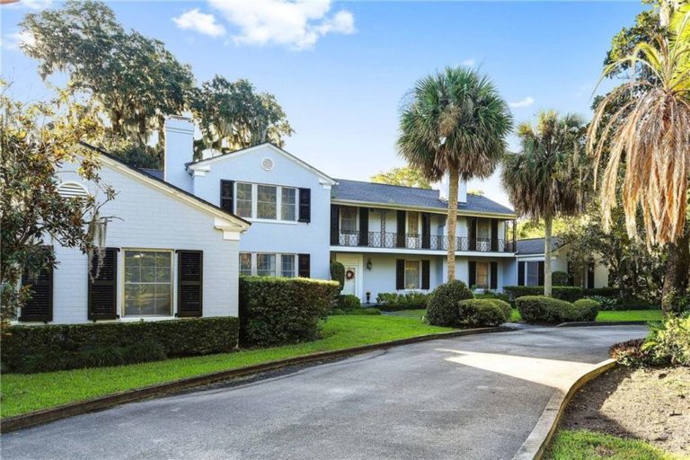 A light blue house sits on green grass with palm trees and a blue sky.