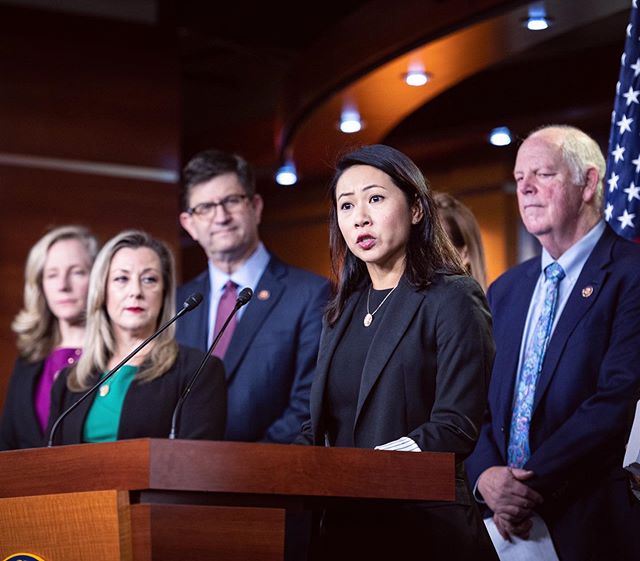 Image shows group of US officials on stage standing behind a podium in a warm wooden room.