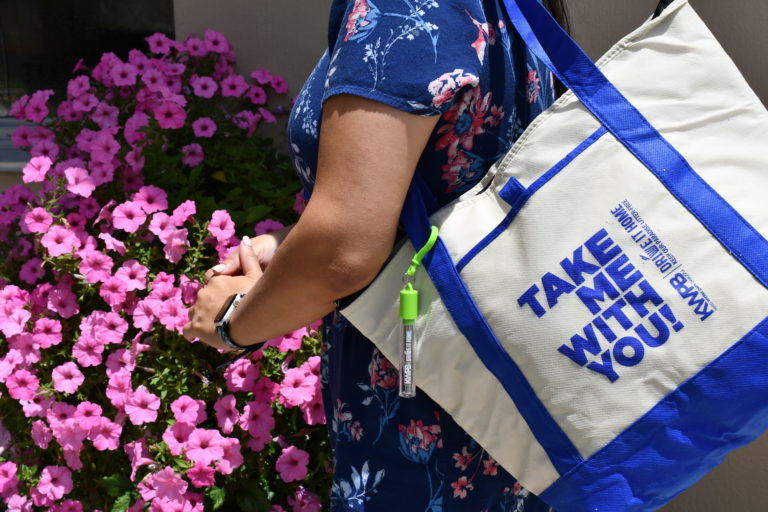 Woman holds large tote bag in front of a bush of flowers.