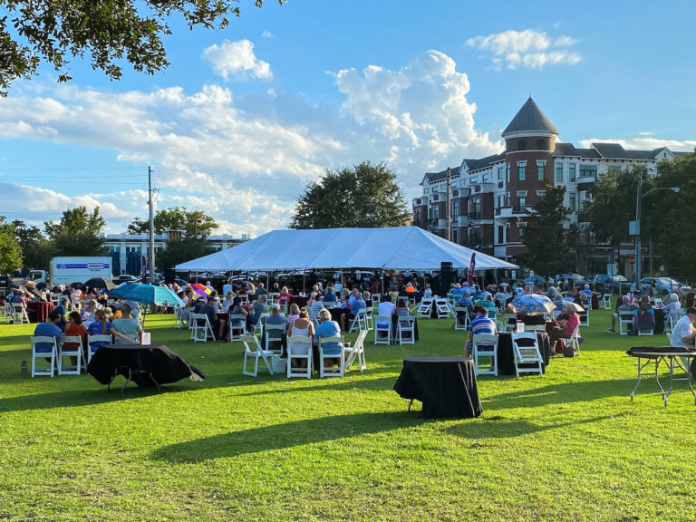 A white tent is shown on a green lawn and blue sky with people sitting at tables.