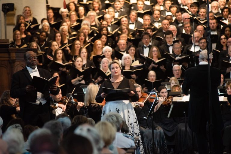Image shows ensemble wearing all black and holding black books to read music from during a concert.