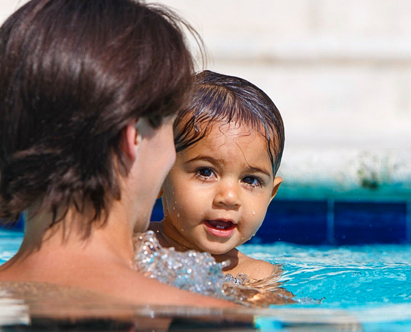 Baby swims in blue pool with teacher in the sunlight.