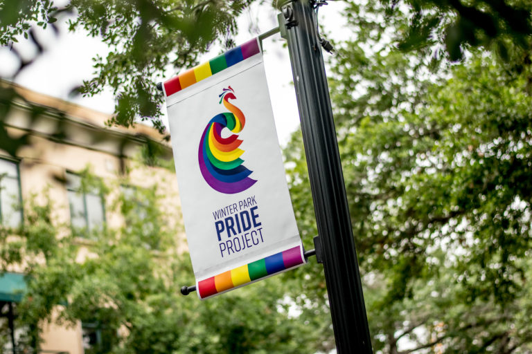 A flag pole shows a Winter Park Pride flag hanging with rainbows in front of green trees.