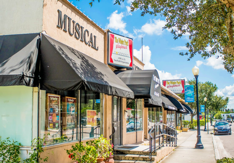Image shows beige building with black awning and sign that says musicals with a blue sky and green bushes