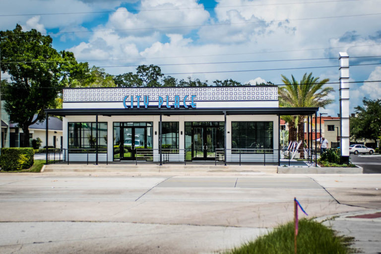 Image shows white building and blue sky with a grey road and green palm trees