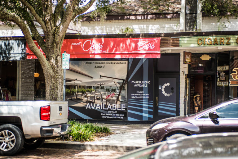 beige building, red awning, black door next to a brown tree with green leaves.